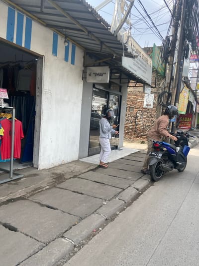 A woman in a grey shirt and pants, wearing a black helmet, stands on the sidewalk in front of a store called "Apple Cloud Store" in Bojongsoang, Indonesia. The store is located at coordinates -6.971 latitude and 107.637 longitude. The woman is holding a small brown purse in her right hand. A man in a brown shirt and grey pants, also wearing a black helmet, is standing on the sidewalk near the woman. He is in the process of getting on a blue motorcycle that is parked on the street. The motorcycle has a black license plate that reads "D 6112 WKB 12-19". The street is paved with asphalt and there is a concrete sidewalk with several cracks. There is a building on the left side of the image and another building on the right side of the image. The building on the left has a blue and white striped awning. The building on the right has a sign that reads "Cloud Store." The sign is in Indonesian.