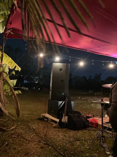 The scene depicts an outdoor event space at night in Murera ward, Kenya. A red fabric canopy is suspended overhead, partially covered by dark palm fronds extending from the upper left. A string of incandescent bulbs runs horizontally across the mid-ground, illuminating the area. Stage equipment is arranged on a grassy surface: a tall, black speaker cabinet labeled "FESTIVA" rests on a shorter, matching speaker cabinet, both positioned on a light-colored wooden pallet. A black electric guitar with a white pickguard leans against the lower speaker, beside a black soft instrument case. To the far right, a partial view of a person's upper torso and arm is visible, standing near a drum kit that includes a visible cymbal on a stand and a drum pedal. In the background, dark tree lines and scattered faint lights suggest an expanse of open ground with other distant activities and figures.