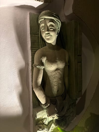 This image, taken in Ao Phai, Thailand, presents a close-up of a stone relief sculpture depicting a smiling, bare-chested female figure integrated into a textured wall. The sculpture portrays the figure with a wrapped head covering, narrow eyes, a small nose, and full lips. Her left arm, bent at the elbow, wears a patterned circular armlet. The torso displays subtle anatomical details and textured skin, while the lower body is draped in a patterned, wrapped garment. Behind the figure, the relief includes a textured panel with vertical striations. The scene is illuminated by artificial light from the lower left, casting prominent shadows on the surrounding light-colored, textured wall to the left and highlighting the sculpture's features. A darker, roughly textured, and curving wall is visible to the right of the sculpture. A faint green light emanates from the bottom edge of the frame, possibly from nearby foliage or a colored light source.