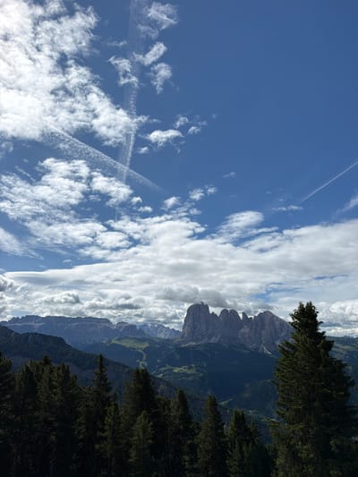 The image captures a panoramic view of the Dolomites mountain range, specifically the Sella Group, as seen from the vicinity of Urtijëi - St. Ulrich in Gröden - Ortisei, Italy. In the foreground, a dense forest of dark green pine trees dominates the lower portion of the frame. Behind the trees, the rugged, jagged peaks of the Sella massif rise majestically against a backdrop of a blue sky filled with scattered white clouds and contrails. The midground reveals rolling hills and valleys, some appearing as green pastures and others covered in more dense forests. The light suggests it is daytime, likely mid-morning or afternoon, with soft shadows indicating diffused sunlight. There are no people or animals visible in the frame. The environment is characterized by its natural, mountainous landscape.