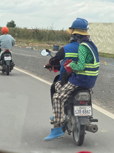 In Krong Ta Khmau, Cambodia, two individuals are riding on a motorcycle, with the passenger in the foreground. The passenger, seated behind the driver, is wearing a blue hard hat layered over a yellow hat and a dark fabric mask covering their head. They are also wearing a bright green long-sleeved shirt with a geometric pattern and a blue safety vest with reflective yellow stripes. Their lower body is covered by plaid pants, and they are wearing bright blue rubber boots. In their hands, they hold a red object. The motorcycle's license plate reads "1JR-8867 PHNOM PENH". In the background, another motorcycle with an orange-helmeted rider is visible. The environment appears to be a roadside with gravel and some vegetation, under a cloudy sky.