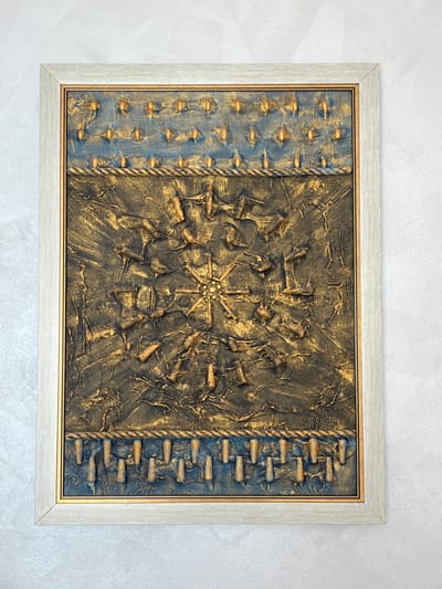 A vertically oriented, framed mixed-media artwork is centrally positioned on a light grey, subtly textured wall. The frame is a light grey/off-white wood with a thin gold inner lining. The artwork's surface is divided into three horizontal sections. The top section features a blue-toned background with multiple rows of small, golden, raised elements, including conical and cross-shaped forms. This section is separated by a horizontal band resembling braided golden rope from the central section. The dominant central section displays a heavily textured, dark blue and gold relief, covered with numerous irregular, golden, three-dimensional shapes. A starburst motif, composed of a central cluster of small, light-colored spherical beads surrounded by radiating golden lines and points, is located at the center of this section. Another golden braided rope-like band separates the central section from the bottom section. The bottom section features a blue-toned background adorned with a horizontal row of golden, downward-pointing conical shapes. The scene is located in Niš, Serbia.