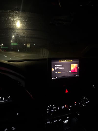 A night scene from inside a car, with a rain-streaked windshield reflecting streetlights. The car's infotainment system displays music information, showing the song "Viernes 13" by Marcos Menchaca. The car's dashboard and climate controls are also visible, illuminated by the internal lights.