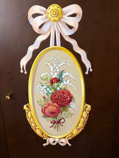 The image depicts a decorative painting on a dark brown wooden surface, possibly a cabinet door. The painting features an oval frame in a muted beige color, surrounded by ornate golden scrollwork at the bottom. At the top, a large, pale pink bow with a golden decorative knot is depicted, with two trailing ribbons that loop downwards. Within the oval frame, a floral arrangement is painted. This arrangement includes two prominent red roses at the bottom, a smaller red rose or bud above them, and delicate white flowers, likely lily of the valley, with green leaves. A small red ribbon is tied around the stems of the roses. The overall style is reminiscent of classical still life paintings with a decorative flourish. The location in Ozeretskoe, Russia, is not discernible from the image itself.