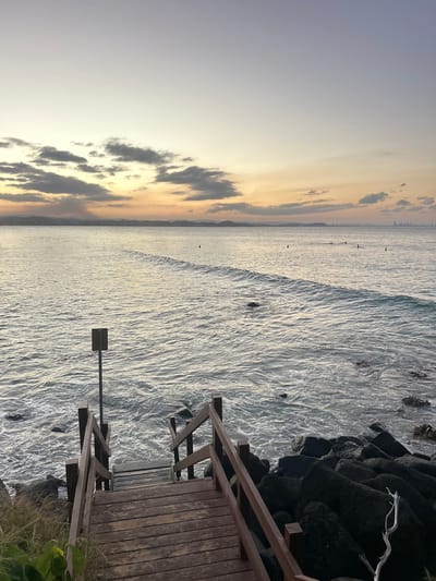 The image depicts a beach scene in Coolangatta, Australia. A wooden staircase descends to the beach. The ocean stretches out, with waves crashing against the shore and rocks. The sky is a gradient of light blues and yellows, indicating either sunrise or sunset. Silhouettes of people can be seen on the horizon in the distance. A signpost stands in the water.