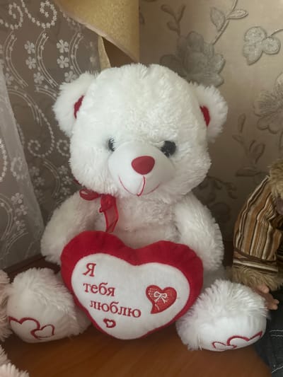 A white teddy bear with red inner ears and a red nose sits on a wooden surface. It is wearing a red ribbon around its neck and holding a red and white heart-shaped pillow. The pillow has Russian text that translates to "I love you" and a small red heart design. The bear's paws also have red heart embroidery. In the background, there is patterned wallpaper and a sheer curtain. To the right of the teddy bear, a portion of another stuffed toy with a striped outfit is visible. The scene is indoors, suggesting it might be a decorative item in a home. The location of Yazykova Pyatina, Russia is not discernible from the image.