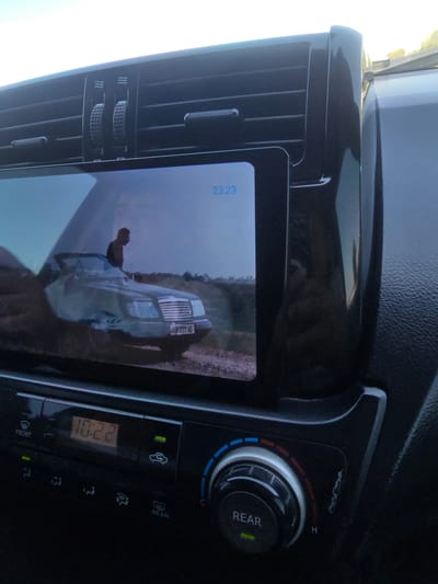 The image shows a car's dashboard with a screen displaying a video. The screen is positioned in the center of the dashboard, above climate control buttons and a digital clock. The clock displays "10:22". The video on the screen shows a person standing next to a convertible car in what appears to be an outdoor setting, possibly a rural area, with a dimly lit sky in the background. The overall environment is the interior of a vehicle, suggesting it is in motion or parked. No specific details about the city of Guzar, Uzbekistan are visible in the image.