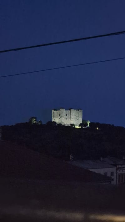 The image, taken at night, shows the Nehaj Tower in Senj, Croatia, illuminated on a hilltop. The tower is a prominent, light-colored stone structure with a crenelated top. It sits atop a dark, tree-covered hill. In the foreground, the outlines of some buildings are visible, also shrouded in darkness. Overhead, two dark power lines stretch horizontally across the deep blue night sky. The overall scene suggests a quiet evening in Senj, with the historic tower standing as a focal point against the night sky.