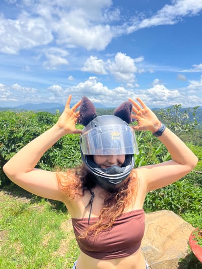 A young woman with curly red hair and cat ears attached to a motorcycle helmet smiles at the camera. She is wearing a brown tube top and holding her hands up in a "peace" sign on either side of her helmet. The background is a lush green landscape with mountains in the distance under a bright blue sky with white clouds. The location is in Sơn Điền, Vietnam.