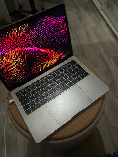The image shows an open laptop with a colorful, abstract wallpaper on its screen. The laptop is silver and appears to be a MacBook Pro, positioned on a round, wooden table with a textured, upholstered edge. The keyboard is visible, featuring a Cyrillic layout. A white charging cable is plugged into the laptop and runs off the edge of the table.
The setting appears to be indoors, with a wooden floor visible in the background. The lighting suggests it could be daytime or evening, with artificial light sources contributing to the overall illumination. There are no people visible in the frame. The overall mood is neutral, focusing on the objects presented.
Visible text on the laptop includes keyboard labels in Cyrillic script, such as "esc", "fn", "control", and "option", along with symbols and numbers.