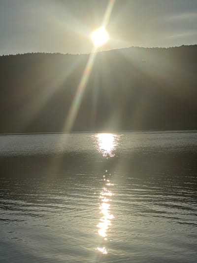 The image depicts a serene natural landscape, likely a lake or a large body of water, with a wooded hill in the background. The sun is low in the sky, casting a bright glare and creating a shimmering reflection on the water's surface. The sky is hazy, with the sun's rays fanning out across the scene, adding a diffused glow. The overall impression is one of tranquility and natural beauty, possibly taken at sunrise or sunset. There are no identifiable structures or people visible. It is not possible to determine the specific location within Sveti Konstantin, Bulgaria from this image alone.