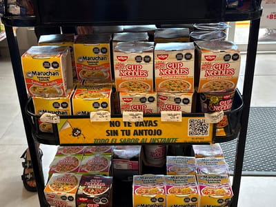 The image displays a retail shelf stocked with various instant noodle products, primarily Maruchan and Nissin Cup Noodles, in different flavors. The products are arranged in rows and stacked in cardboard boxes and individual cups. Some boxes are open, revealing the contents. Prominent yellow signage with black text reads "NO TE VAYAS SIN TU ANTOJO TODO EL SABOR AL INSTANTE." Below this, a QR code is visible. The shelf unit appears to be made of black metal. In the foreground, a person's legs and feet wearing black sandals are visible, suggesting they are a shopper. The background is slightly blurred but shows a store interior with other merchandise and a textured floor mat. The image captures a commercial setting, likely a convenience store or supermarket aisle, in San Juan Tejaluca, Mexico. The specific details of the products and the Spanish signage confirm the location.