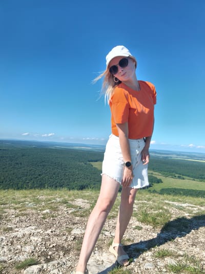 A young blonde woman wearing a white baseball cap, black sunglasses, an orange crop top, and a white denim skirt stands on a rocky outcrop overlooking a vast green valley under a clear blue sky. She is leaning to her left with her head tilted and is looking towards the camera. Her right arm is bent at the elbow, and her left arm hangs at her side. She is wearing beige wedge sandals and a watch on her left wrist. The background shows a dense forest that stretches to the horizon, with patches of lighter green fields and distant hills. The location is identified as Shikhan, Russia.