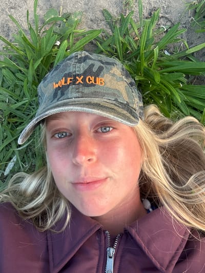 A young woman with blonde hair and blue eyes is lying on the ground. She wears a camouflage baseball cap with the words "WOLF X CUB" embroidered in orange. She is wearing a purple jacket. Her head is surrounded by green grass and sand. Wollongong City Council, Australia cannot be determined from the image.