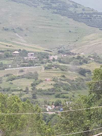 This image depicts a rural village nestled within a valley in Bazum, Armenia. The scene is a wide shot, showing a landscape dominated by rolling, green hills and mountains. The lower portion of the image reveals a village with scattered houses and buildings. A dirt road or path winds its way through the village. Several trees and shrubs are visible, some in the foreground and others dotting the hillsides. A light-colored fence runs horizontally across the midground, separating different sections of the village or property. In the distance, the landscape is covered in lush vegetation, with some areas appearing more sparsely vegetated. The sky is overcast, suggesting a cloudy or possibly rainy day. There are no people visible in the image, and the overall impression is one of quiet, rural life. The time of day is indeterminate due to the cloud cover, but the lighting suggests it is daytime. The presence of houses and a road indicates a settled community. There is no visible text in the image.