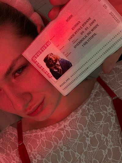 The image features a person holding what appears to be a humorous, mock passport labeled "MOONLAND" with a photo of a cat wearing a suit. The passport contains some Polish text including "BOBR" and phrases like "JAKIE BYDLĘ JEBANE" and "SPIERDOLIŁ DO WODY I SIĘ UTOPIŁ", as well as the birth date "09.01.2009" and the date "202020". The scene is illuminated with a red light, and the person holding the passport is partially visible, lying down and wearing a white lace top with red straps.