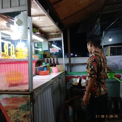 The image shows a man wearing a floral shirt, standing in front of a food cart in Kecamatan Legok, Indonesia. The man is holding a spatula and appears to be cooking food in a wok on a small stove. The food cart has a metal counter with a large red plastic basket containing food. The cart also has a sign that says “TUTUP CLOSED.” There are several glass bottles of what appear to be sauces or beverages, along with other kitchen utensils. The image appears to have been taken at night, as the lighting is dim and the sky is dark. The environment is cluttered with various objects, including a green bucket and a blue box. The man is looking down at the wok, focused on his task. The location of the food cart is based on the provided coordinates (-6.273, 106.55), which corresponds to a street in Kecamatan Legok, Indonesia.