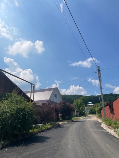 The image shows a residential street in a village. The sky is blue with scattered white clouds. A paved road runs through the center of the image, curving to the right. On the left side of the road, there is a house with a metal roof, a green bush, and a wooden structure. On the right side, there is a red corrugated metal fence and a utility pole with wires. In the background, there are more houses and a hill covered in trees. The scene appears to be a sunny day.