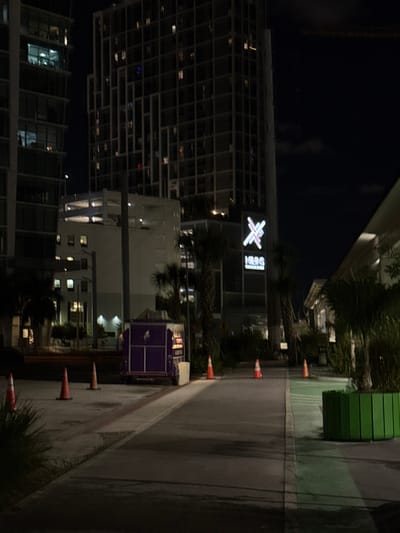 A nighttime urban view presents the "X Miami" building with its illuminated logo at the top. Several traffic cones line a wet, dark sidewalk beside palm trees. A green dumpster sits further along the path. Nearby, a purple equipment box appears against the backdrop of other buildings. The scene is illuminated by artificial lighting, and a tall building with a distinct pattern of lighted windows dominates the left side of the image.