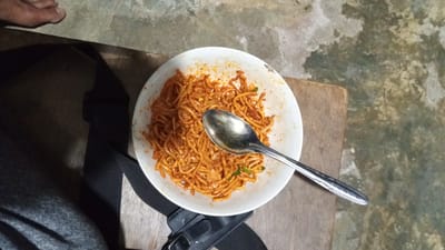 A close-up, overhead shot captures a white ceramic bowl filled with stir-fried noodles, seasoned with red chili powder and what appear to be green chilies. A metallic spoon rests inside the bowl, partially submerged in the noodles. The bowl sits on a rustic wooden surface, which is adjacent to a rough, speckled concrete floor. In the upper left corner of the frame, a portion of a person's foot is visible, suggesting the presence of an individual in the scene. A dark strap, possibly from a bag, runs diagonally across the lower portion of the image, obscuring part of the wooden surface and a person's gray clothing. The overall lighting is dim, casting shadows and giving the scene a somewhat muted appearance. The image does not provide specific visual cues to definitively pinpoint its location within Cotgadong, Indonesia, beyond the general setting of a domestic or informal eating environment.