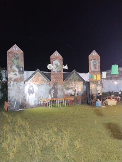 The image depicts an outdoor scene in Jetis, Indonesia, at night. The dominant feature is a makeshift castle-like structure with painted images of various ghoulish or monstrous figures, including skeletons, pale women with long dark hair, and other creatures. The structure is built of brick-patterned surfaces and adorned with triangular peaks. Attached to it are loudspeakers.
In front of the structure is a grassy area. To the right of the castle structure, two men are sitting, appearing to be resting. Near them are a blue barrel, a cooler, and a food vendor stand. The backdrop includes a concrete wall with graffiti and some hanging lanterns. The lighting is dim, with the structure and immediate area being lit.