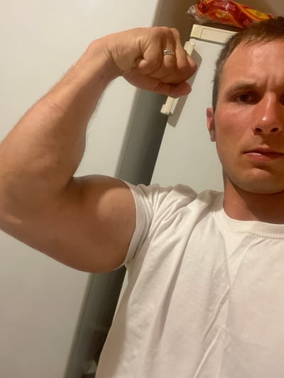 The image shows a close-up of a man's bicep as he flexes his arm. He is wearing a white t-shirt. His arm is tanned and appears muscular. The background is out of focus but appears to be an indoor setting with a white wall and possibly a refrigerator. The image is taken from a low angle, looking up at the man's arm and face. There is no information in the image to suggest it is located in the city of Турынскі сельскі Савет, Belarus.