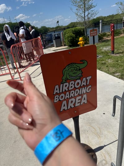 The image depicts a person standing in line for an airboat ride in Fort Lauderdale, Florida. The sign indicates the "Airboat Boarding Area" with an alligator cartoon. A queue with people waiting is visible behind a red metal barrier. The person has a blue wristband with black lettering and neatly manicured nails on their hand that is in the foreground. In the background, there is a green lawn, some trees, a red fire hydrant, and a yellow fire hydrant. The scene is outdoors, on a sunny day.
