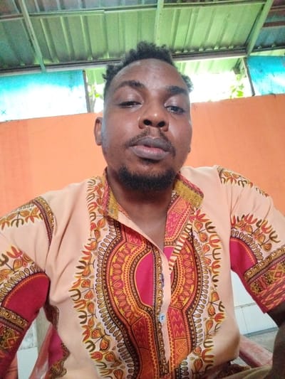 In an outdoor, open-air setting possibly under a shelter, a young Black man is pictured from the chest up. He has dark, curly hair, a goatee, and is looking directly at the camera with a neutral expression. He is wearing a colorful dashiki shirt with intricate geometric and floral patterns in shades of orange, red, yellow, and brown. The background consists of an orange wall, a green corrugated metal roof supported by a metal structure, and some foliage visible through an opening. The lighting suggests it is daytime. The location is not identifiable as Pekanbaru, Indonesia.