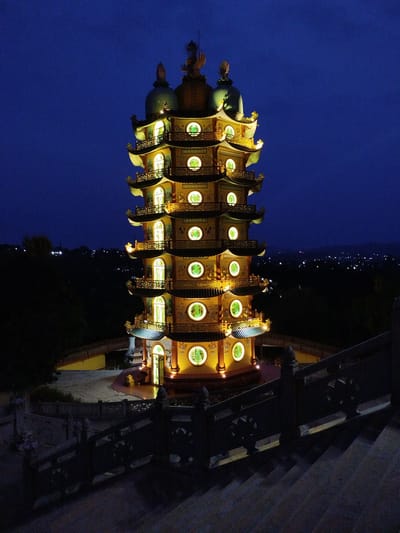 The image depicts the illuminated Bửu Long Temple in Phú An, Vietnam, at night. The multi-tiered pagoda is centrally positioned, its golden facade contrasting against the dark blue sky. Each level features circular windows emitting a soft, warm glow, and traditional architectural details like curved eaves and decorative embellishments. The pagoda sits atop a paved platform enclosed by railings. A flight of stairs with intricately patterned stone railings leads towards the structure. The background reveals a dark landscape with scattered city lights in the distance, suggesting a vantage point offering a view of the surrounding area. The foreground is defined by the descending staircase, subtly indicating the viewer's perspective from a higher elevation.