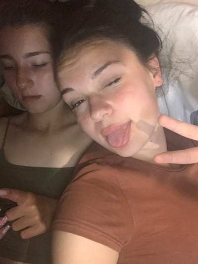 The image shows a close-up, overhead view of two people, likely young women, lying down. One person is in the foreground, with her face angled towards the camera. She has dark hair and is sticking her tongue out, with two fingers extended in a peace sign gesture, suggesting a playful or lighthearted mood. Her skin appears fair, and she has dark eyebrows. The second person is positioned below the first, with her eyes closed and her face partially obscured. Her hair is lighter than the first person's. Both individuals are wearing casual clothing, with the person in the foreground in a brown shirt and the person underneath in a green top. The background appears to be a bed with white sheets. The lighting is dim, suggesting an indoor setting and possibly nighttime or a poorly lit room. There are no visible text elements in the image. The overall impression is one of intimacy and casual interaction.
