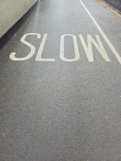 The image shows the word "SLOW" stenciled in white paint on an asphalt surface, likely a road or pathway. The letters are large and clearly visible. The asphalt appears to be well-maintained, with some small debris and leaves scattered on its surface. To the left of the painted word, there is a low, light-colored concrete wall that runs parallel to the pathway. To the right, a white line is painted on the asphalt, demarcating a lane or edge. In the background, a dark metal fence is visible, with some overgrown vegetation behind it. The scene suggests an outdoor urban or suburban environment. The specific location in Stockton-on-Tees is not identifiable from the visual information alone. There are no people or animals present in the image.