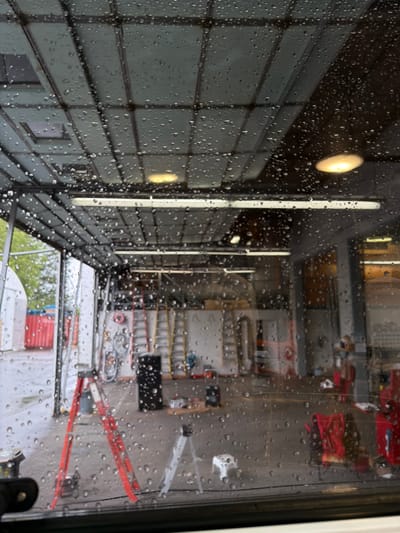 The image shows a reflection on a window covered in raindrops. The reflection appears to be of an indoor setting, possibly a garage or workshop, with an outdoor scene visible through a large opening.

Inside the reflected space, various objects are visible, including what appears to be industrial equipment, shelving units, and red ladders leaning against a wall. The lighting suggests it is daytime, with a prominent light fixture reflecting on the left side of the image. The overall impression of the interior is organized and functional, with tools and equipment neatly arranged.

The outdoor scene visible through the opening shows greenery, suggesting trees or bushes, and some red containers. The presence of rain on the window indicates it is a wet or rainy day. The image is taken from a perspective looking through a window, so the subjects and their details are observed as reflections. No people are directly visible, but the presence of equipment suggests human activity might be ongoing or has recently occurred.

The location is specified as Delta, Canada. The time of day is likely daytime, given the artificial light and the visible outdoor scene. The weather is clearly rainy. The overall mood conveyed is somewhat muted due to the rain and reflections, but the glimpse of the workshop suggests a busy or productive environment. No legible text is visible in the image.