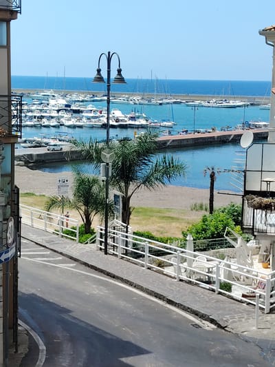The image shows a view of a marina with numerous boats docked, a clear blue sky, and the sea. In the foreground, a street curves away from the viewer, bordered by a sidewalk and a white railing. Beyond the railing, there is a patch of sand, followed by green grass and trees. A person is visible standing on the sidewalk, wearing shorts and a light-colored top. Signs are posted near the sidewalk, one with a "30" speed limit and an arrow. A lamppost with two lights stands near the railing. The location is the port of Agropoli, Italy, a coastal town in the Campania region.