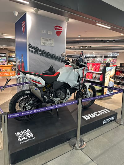 The image shows a white Ducati DesertX motorcycle displayed in an indoor location, likely a shopping center in Pudahuel, Chile, as indicated by "ducatichile.cl" on the display. The motorcycle is positioned on a black platform with the Ducati logo and surrounded by stanchions with purple "despegar" branded straps. The backdrop features Ducati branding and a depiction of a road, with other commercial advertisements visible in the background. Products are visible on shelves, suggesting the area is a retail space. Signage indicates the sale of alcoholic beverages.