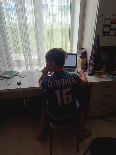 A boy is sitting at a desk in a room, likely in an apartment, in the city of Closed administrative–territorial formation "Mezhgorye", Russia. He is wearing a blue athletic jersey with the name "СЕЛЕЗНЕВ" and the number "16" on the back. He is looking at a laptop on the desk and his hand is near his mouth, suggesting he may be thinking or concentrating. The desk is cluttered with various items including a notebook, papers, and a pair of wireless earbuds. A window is behind the desk with sheer curtains and a view of an apartment building and trees outside. A cabinet is to the right of the desk with some items on top of it. The floor is visible and partially covered by a black bag.