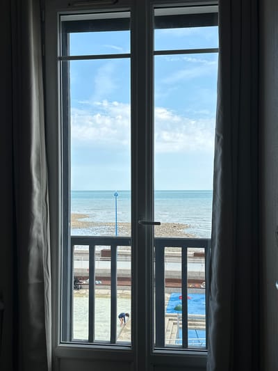 The image shows a view through a French window looking out onto the sea. The window is divided into two vertical panes, with a curtain drawn back on either side. The sea is calm and a light blue, meeting a pebble beach in the foreground. Beyond the beach is a railing, and then an area with a blue mat, some toys, and a child playing in shallow water. There is also a stroller visible. The sky is blue with some white clouds. The setting appears to be a beachside resort or promenade. The presence of the blue mat and toys suggests a children's play area, possibly a splash pad or shallow pool, situated between the promenade and the beach. A blue pole with a white top is visible on the pebble beach. The overall impression is a bright, sunny day at the seaside.