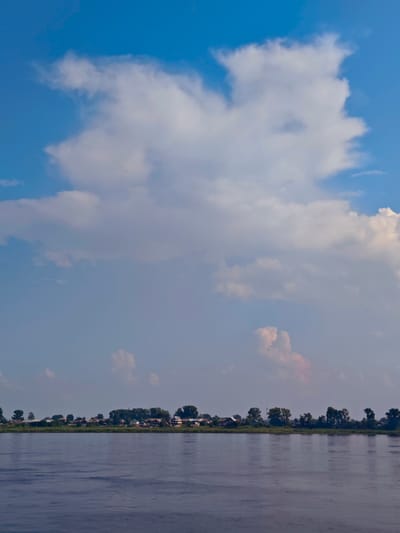A wide, dark blue river occupies the bottom half of the image. On the far bank, a line of trees and houses form a settlement. The houses are small, with peaked roofs and are situated close together. Behind the settlement, the trees are dense and green. The sky above is a clear, bright blue, with large, fluffy white clouds scattered across it. One large cloud formation dominates the upper left portion of the sky, with softer, pinkish clouds visible lower down and to the right. The image appears to be a landscape view of a rural settlement on the banks of a river, possibly in Yeniseysk, Russia, given the typical scenery of the region. There are no people visible in the image.