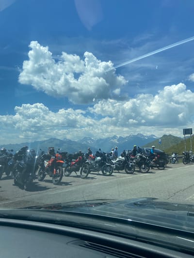The image captures a scenic view of a mountain pass, likely in the Italian Alps, with a group of motorcycles parked by the roadside. In the foreground, the dashboard of a vehicle suggests the photo was taken from inside a car. The main subjects are a collection of dual-sport and adventure motorcycles, varying in color, parked in a line along the asphalt road. Some motorcycles are orange, red, black, and white. Several individuals are visible, some standing near the bikes, and others are seated on them, wearing helmets and motorcycle gear. The backdrop features majestic snow-capped mountains under a bright blue sky adorned with fluffy white clouds. The environment appears to be a high-altitude location, possibly a popular stop for motorcyclists. A signpost can be seen on the right side of the image, and a few more signs further up the hill, suggesting it is a marked viewpoint or pass. The overall scene conveys a sense of adventure and appreciation for the mountainous landscape. The location is identified as St. Leonhard in Passeier - San Leonardo in Passiria, Italy.