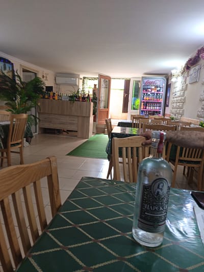 The image depicts the interior of a restaurant or cafe in Derbent, Russia. In the foreground, a glass bottle of alcohol, possibly vodka, with a label featuring Cyrillic text and a portrait, rests on a table covered with a green tablecloth patterned with a diamond grid. The bottle is partially filled with a clear liquid. The table is surrounded by wooden chairs with slatted backs.
In the mid-ground, several more wooden tables and chairs are visible, some covered with similar green tablecloths. Beyond the tables, a wooden counter or bar is present, stocked with numerous bottles of beverages. A Coca-Cola branded refrigerator unit is also prominent, filled with drinks. The background shows a doorway leading outside, revealing greenery and daylight. An air conditioning unit is mounted on the wall above the counter. Decorative elements, such as garlands of tinsel, are visible along the ceiling. The flooring consists of light-colored square tiles. The overall impression is that of a casual dining establishment.