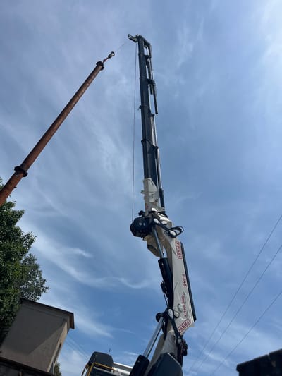 An outdoor, low-angle, medium shot captures a construction scene against a blue, partly cloudy sky in Castiglione delle Stiviere, Italy. The primary focus is on a white and black FASSI F54-5 truck-mounted crane's articulated arm, which is extended upwards. The crane's branding, "FASSI" and "OMC GRU," is visible on its white segments. A thick, rusty, brown pipe is suspended from the crane by a cable and a metal clamp. The pipe angles diagonally from the left to the center of the frame. In the lower left, the green foliage of a tree and the upper edge of a small, light brown, boxy structure are visible. In the lower right corner, a portion of a white vehicle, possibly the truck on which the crane is mounted, is seen, along with a few dark overhead power lines. No people are visible in the image. The scene appears to depict a pipe being lifted or moved by the crane.