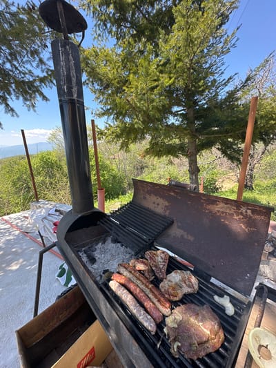 The image depicts a barbecue grill in an outdoor setting, possibly Lyubovka, Bulgaria, considering the prompt's request. On the grill, there are various meats, including sausages and what appears to be steaks. Some pieces of onion can also be seen. A chimney extends from the grill, and the surrounding area includes trees and a clear sky. The grill sits on a stand, and there are other objects nearby, such as a box and what might be a plate or container. The scene suggests an outdoor grilling event.