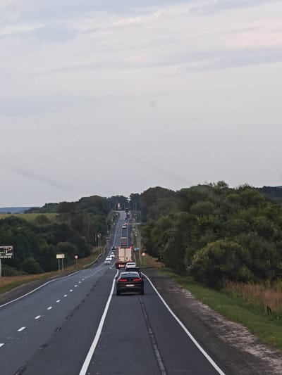 The image depicts a highway in Kizhevatovo, Russia, with a line of vehicles traveling uphill. The road has two lanes in each direction, separated by white lines. On the right side of the road, there is a grassy embankment with vegetation. On the left side, there are fields and a dense forest. The sky is overcast with gray clouds.
In the foreground, a black SUV is driving on the right lane. Further up the road, there are several other cars and trucks. A large white truck is visible near the top of the hill. The overall scene suggests a typical highway scene in a rural or semi-rural area.