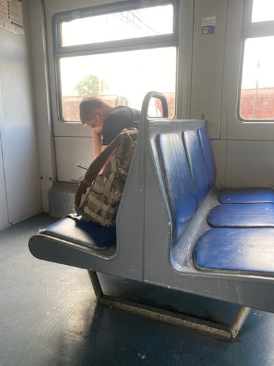 The image captures the interior of a train car, possibly in Romodan, Ukraine, with a young man seated on the left side, facing the window. He appears to be looking down, possibly at a mobile device held in his hands, with his head resting on his hand. He is wearing a black t-shirt and has earbuds in his ears. A camouflage backpack is placed on the seat next to him. The train car features blue upholstered seats and a grey partition. Natural light streams in from the windows, illuminating the scene and casting shadows on the floor. The environment suggests a mode of public transportation, likely a train, given the seating arrangement and windows. The overall impression is one of quiet travel, with the passenger engaged in personal activity.