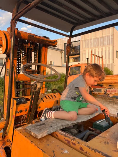 A young boy is sitting on a piece of wood inside the engine compartment of an orange, rusty forklift. He is holding a black object. The background includes a building with large windows and another orange vehicle, potentially a truck. The location is Pobrdje, Montenegro.