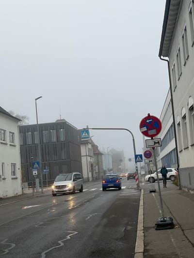 The image depicts a street scene in Tuttlingen, Germany, under foggy and overcast conditions, with wet road surfaces.

In the foreground, on the right, a gray pavement sidewalk features a temporary street sign assembly. This assembly includes a red circular sign with a blue diagonal bar and a red 'X' (indicating "No stopping or parking") pointing left, augmented by a lower sign depicting a vehicle being towed and another red circular sign with a blue background and a single red diagonal bar (indicating "No parking"), also pointing left. These signs are supported by a metal pole anchored by two black plastic bases.

The two-lane asphalt road ahead is wet and reflects ambient light, showing faint lane markings. A silver Mercedes-Benz van/minibus with its headlights illuminated is moving towards the viewer in the left lane. Following it in the right lane, a blue Volkswagen Golf is driving away, displaying its red taillights. Further down the street, multiple vehicles are visible as indistinct shapes with illuminated headlights and taillights, partially obscured by the dense fog.

On the left side of the street, a multi-story white building with numerous windows stands, adjacent to a more modern multi-story building featuring a dark gray, vertically ribbed facade and large windows. In front of this modern building, a street pole holds a blue square sign with a white pedestrian crossing triangle and a white directional arrow.

Spanning overhead across the road, a metal gantry structure supports a pedestrian crossing sign (blue square with a white triangle and walking person icon). A tall, dark street light is positioned near the modern building on the left.

On the right side of the street, further down, a person is walking on the sidewalk away from the viewer, wearing a dark green jacket. A white SUV is parked on the right, partially visible behind the pedestrian. The background is heavily obscured by thick, uniform gray fog, rendering distant buildings and features indistinct.