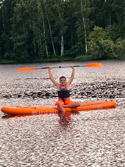 A young person wearing a life vest and swim goggles is seated on an orange inflatable paddleboard on a body of water. The person is holding an orange paddle above their head with their right arm raised. The paddleboard has "FUSION" written on it. The water is reflective, and in the background, a dense forest of green trees is visible against a cloudy sky. The location is in Vecumnieki, Latvia.