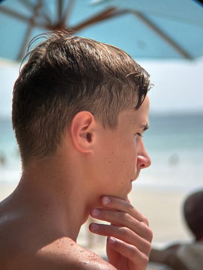 This is a close-up, outdoor shot of a young person's head and upper body. The subject is looking upwards and to the right, with their hand resting on their chin and neck area. Their hair appears wet and slicked back, suggesting recent exposure to water. Droplets of water are visible on their skin, particularly on their neck and shoulder. The lighting suggests it is daytime, with bright sunlight casting shadows. The background is out of focus, with areas of bright light and a soft blue hue, possibly indicating a bright sky or water. The overall impression is one of relaxation and perhaps contemplation. There is no visible text in the image. The location context of Dong Ninh Hoa, Vietnam, does not provide further clues about the specific activity or setting.