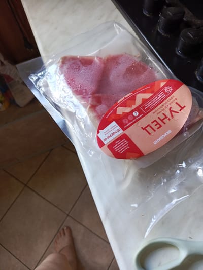 The image shows a package of meat, likely vacuum-sealed, lying on a tiled surface next to a pair of scissors. The packaging has a red and pink label with Russian text. Part of a person's foot and leg is visible in the lower left corner of the image, suggesting they are standing or sitting near the meat. The environment appears to be indoors, with the tiled floor and a countertop or table visible. There are also some dark, cylindrical objects on a black surface in the background, possibly part of a stove or appliance. No specific location in Alexandrovskaya, Russia can be determined from the visual information alone.