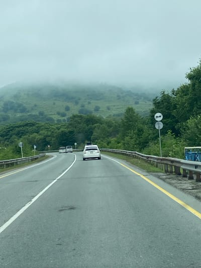 The image depicts a highway scene in Bamburovo, Russia, characterized by a dense fog obscuring the distant hills. The road, with a white dividing line and a yellow edge line, curves to the left. Several vehicles are visible: a white SUV in the foreground, and further ahead, two white sedans. The surrounding environment consists of lush green trees and vegetation, with a guardrail lining the right side of the road. Two traffic signs are also present: one with two cars stacked vertically, indicating a possible overtaking or passing restriction, and another circular sign below it. The overall atmosphere is muted due to the overcast and foggy conditions.