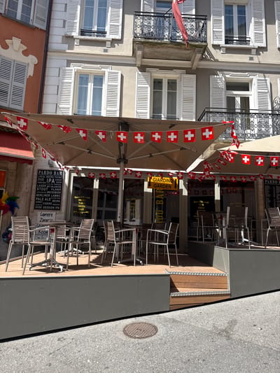 This is a medium shot, rotated image of an outdoor cafe or bar area in Locarno, Switzerland. The scene is set on a sunny day, indicated by the strong shadows. The establishment, named "Pardo Bar," is decorated with numerous small Swiss flags strung along lines, suggesting a festive or patriotic atmosphere.

The foreground features several tables and chairs, some stacked. A large, light-colored awning provides shade over part of the seating area. Visible text on signs includes "Pardo Bar," a menu listing various drinks like "IPA," "Stout," and "Pale Ale," and "Giardino Garden." Another sign lists "Shisha," "Table," "Soccer," and "Dart," indicating the services offered. Window signage also displays "Vino," "Birra," and "More."

Behind the cafe, a building with light-colored walls and white shutters on multiple windows is visible. The architecture suggests a European style. The street is visible in the background on the right, with a manhole cover on the asphalt. The overall impression is a casual outdoor dining and drinking establishment ready to serve customers. No people are actively depicted in the image.